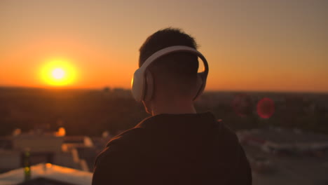 A-man-walks-on-the-roof-at-sunset-with-headphones-looking-at-the-city-from-the-height-of-a-skyscraper-at-sunset.-Relax-while-listening-to-music.-Enjoy-a-beautiful-view-of-the-city-at-sunset-from-the-roof-with-headphones